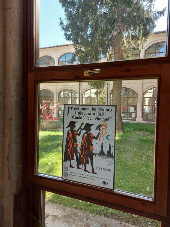 Cartel en ventana: Dos figuras disfrazadas con paisaje urbano, probablemente un anuncio de un evento. Patio exterior.