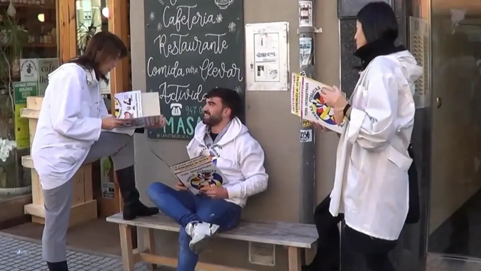 Tres personas afuera de un café: dos sosteniendo libros, la otra sentada, leyendo un libro, cerca de un menú en una pizarra.
