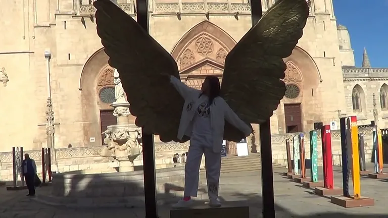 Persona con los brazos extendidos, posando con una escultura de alas de ángel frente a una iglesia.