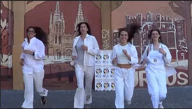 Cuatro mujeres corriendo, sonrientes, vestidas con ropa blanca, frente a un mural con un edificio.