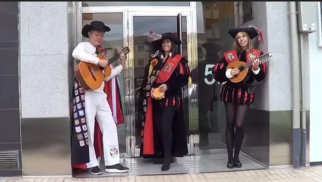 Tres personas con trajes renacentistas tocan guitarras afuera de un edificio con una puerta de vidrio; día soleado.