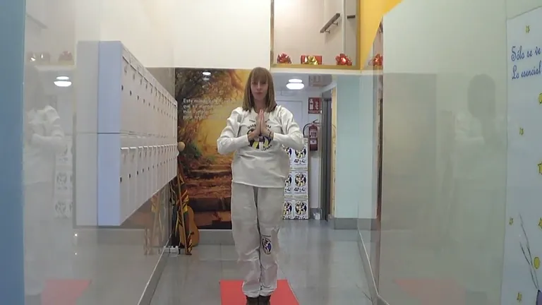 Mujer con traje de yoga blanco, con las manos en oración, haciendo equilibrio sobre una pierna sobre una colchoneta roja en el interior.
