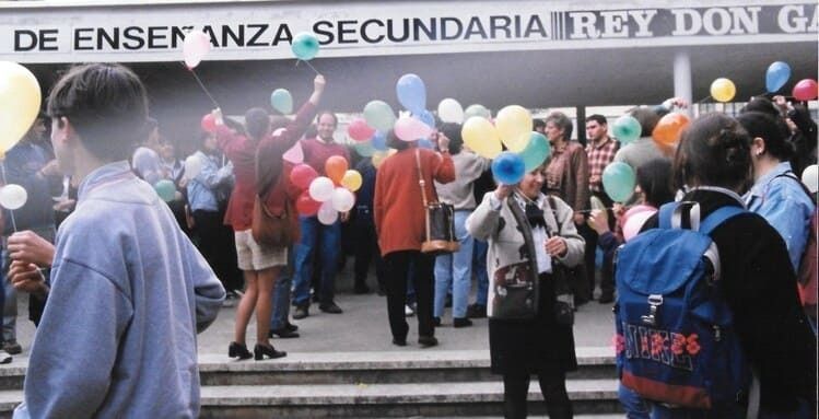 Personas con globos afuera de un instituto. Ambiente alegre; colores variados; celebrando.