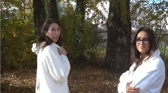 Dos mujeres con sudaderas blancas están paradas en un parque con árboles y hojas caídas, mirando a la cámara.