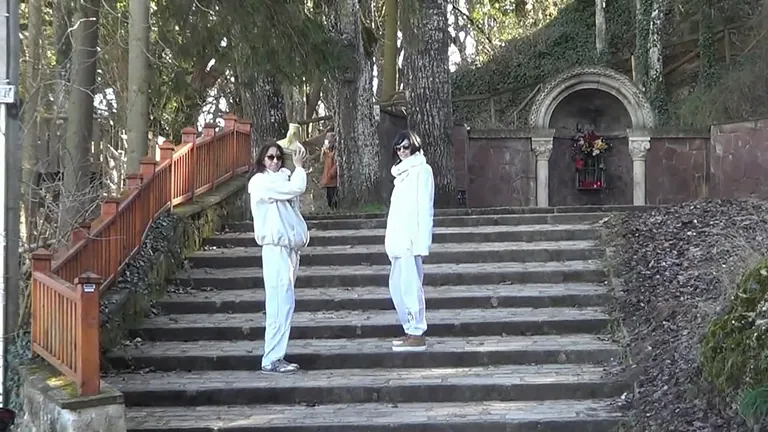 Dos personas vestidas de blanco de pie sobre unos escalones de piedra al aire libre; nicho de piedra con corona de flores al fondo.