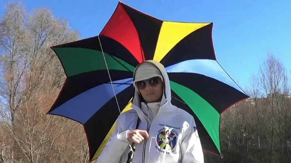 Persona que viste una chaqueta blanca y gafas de sol, sosteniendo un paraguas grande y colorido, de pie al aire libre.