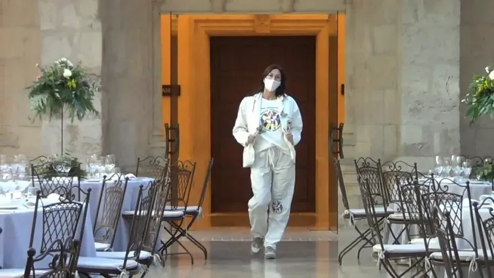 Mujer con una máscara facial, caminando hacia la cámara en un comedor formal con mesas preparadas para una reunión.