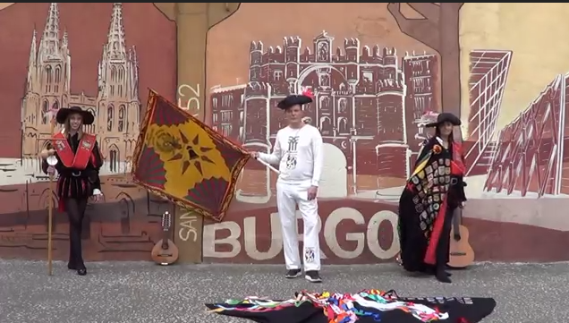 Personas disfrazadas con una bandera en Burgos, España, frente a un mural; uno sostiene una bandera con un diseño de sol.