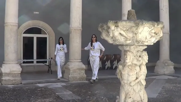 Dos personas vestidas de blanco caminan hacia la cámara cerca de una fuente clásica y columnas.