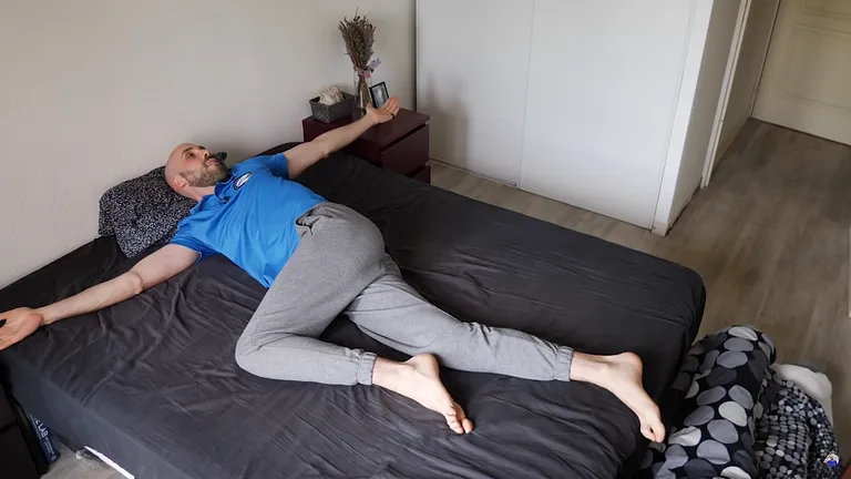 Hombre acostado en la cama, con los brazos extendidos, las piernas flexionadas y el torso girado. Pantalón de chándal gris, camisa azul, posición neutra.