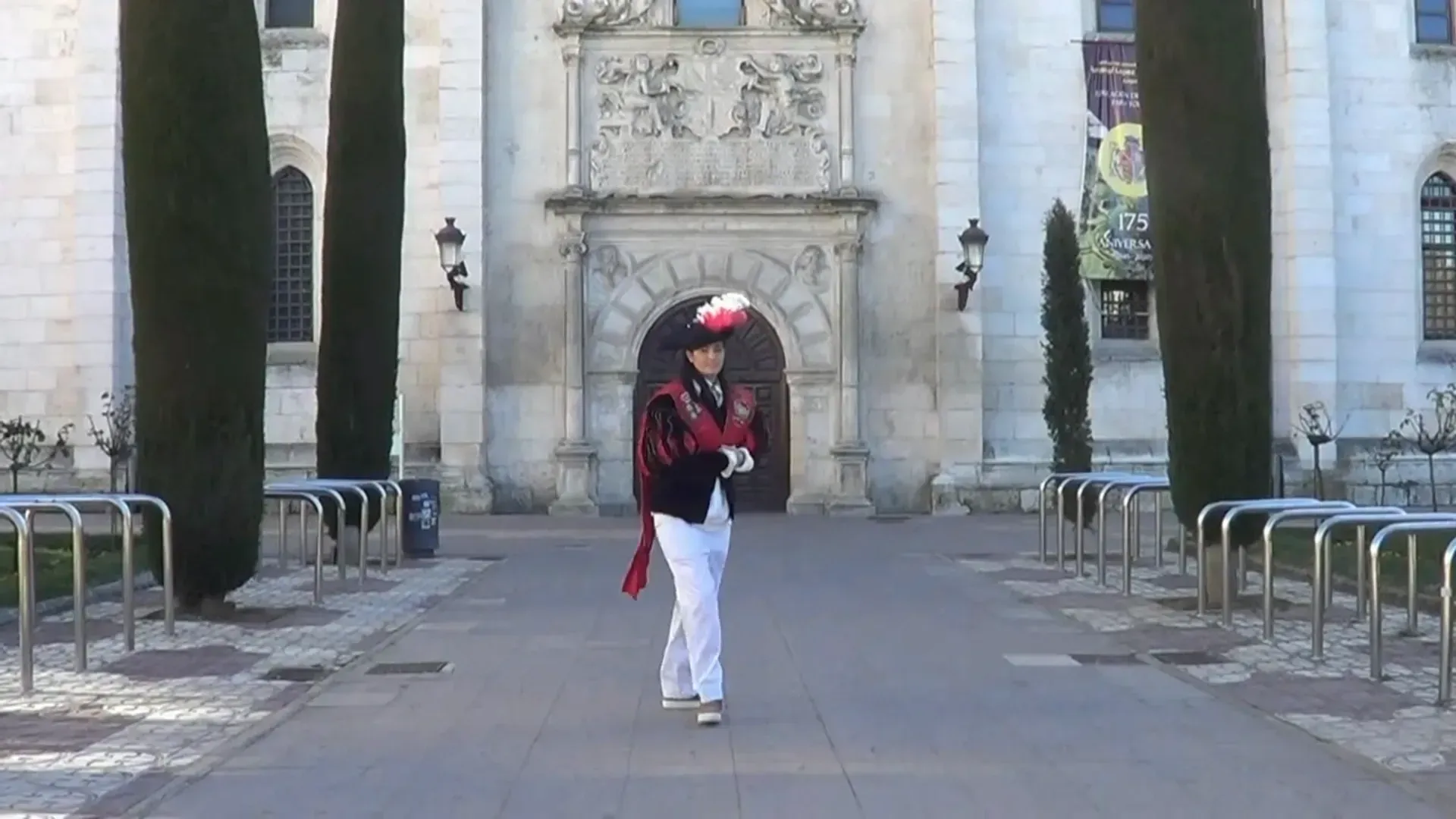 Persona con atuendo ornamentado caminando hacia la cámara frente a un edificio detallado con columnas.