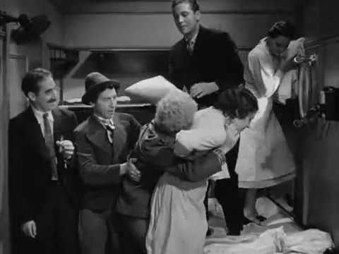 Escena en blanco y negro: Varias personas rodean y abrazan a una mujer en una habitación; un hombre observa.