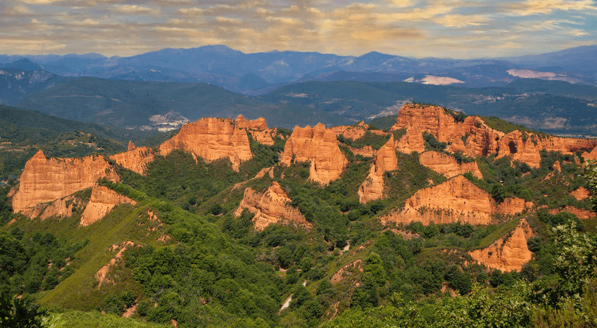 Formaciones rocosas anaranjadas que se elevan sobre árboles verdes, con montañas en la distancia.