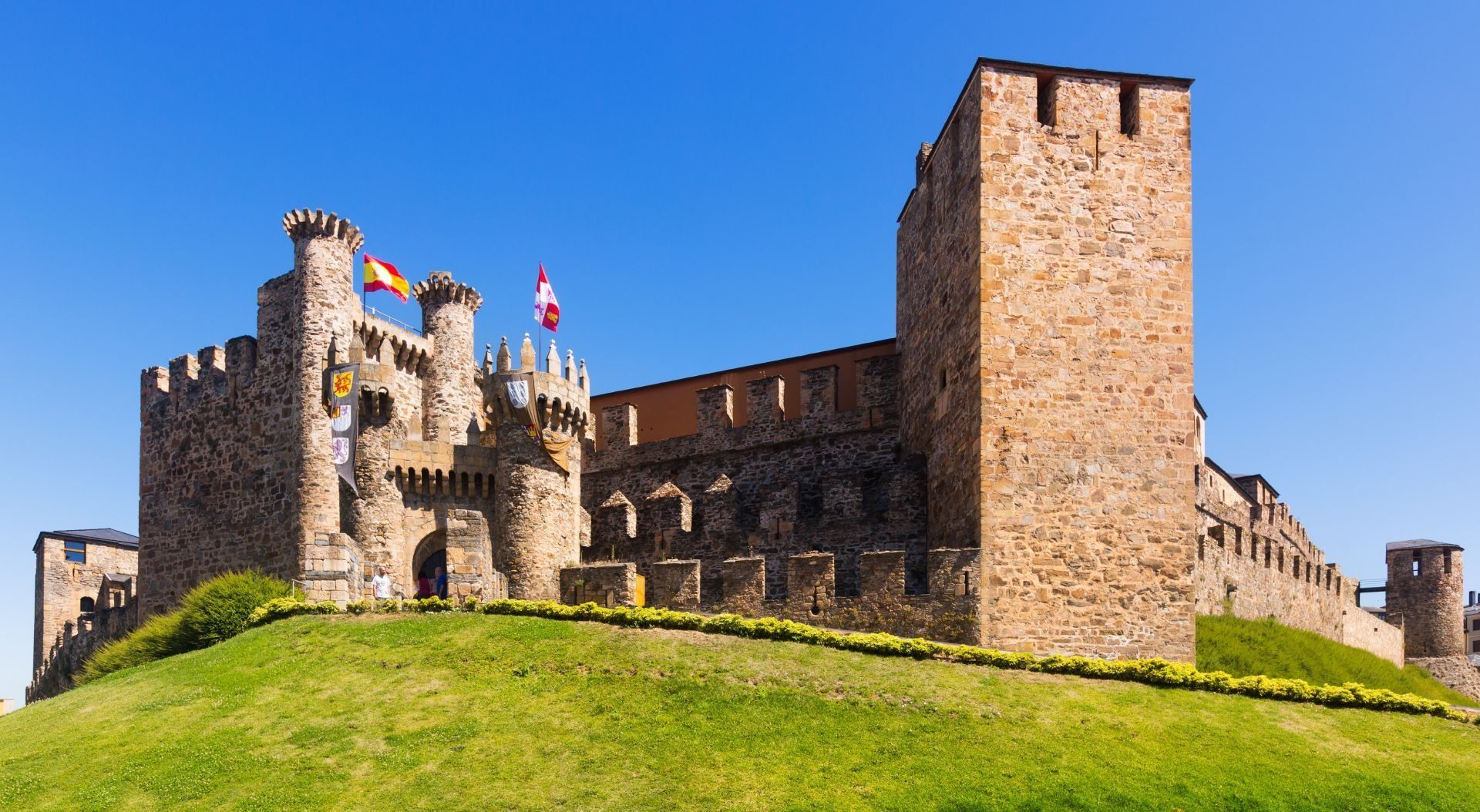 Castillo de piedra con torres y almenas en lo alto de una colina verde bajo un cielo azul claro.