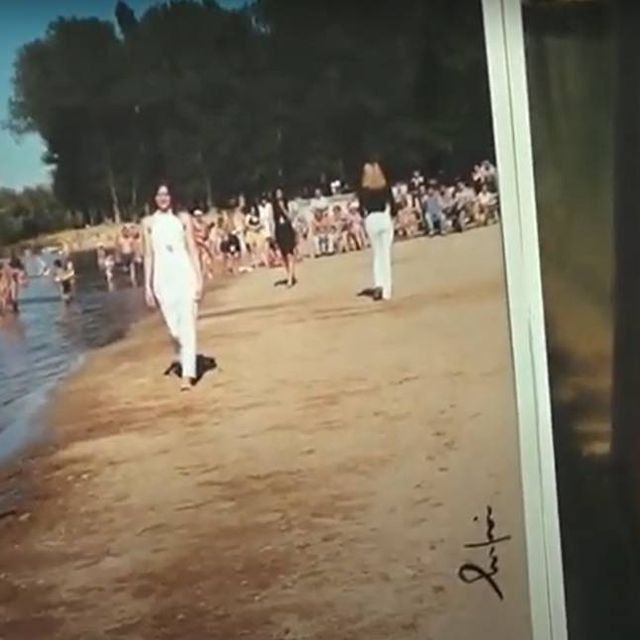 Escena de playa: Gente cerca del agua y en la arena. Dos mujeres vestidas de blanco pasan caminando.