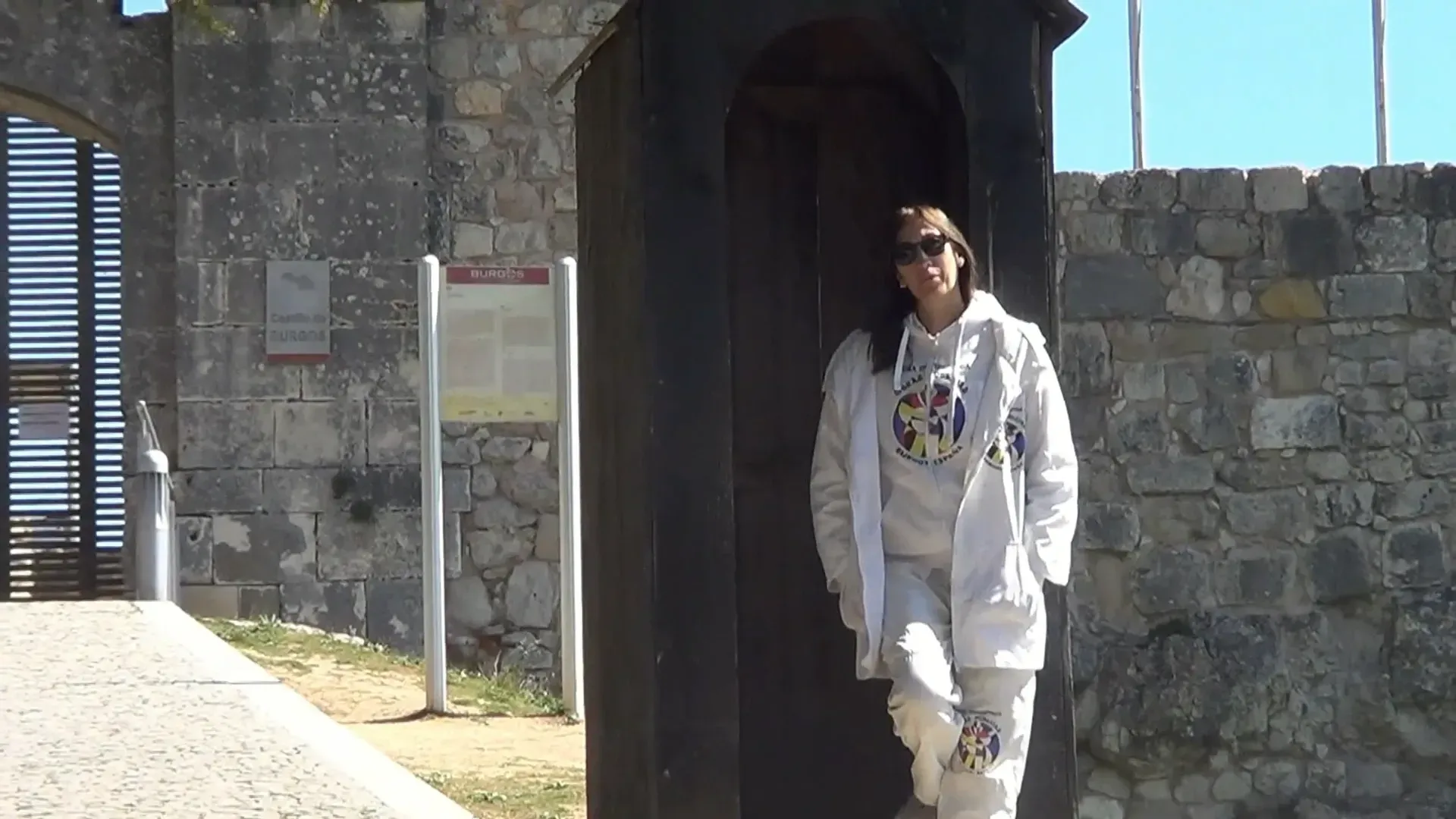 Mujer vestida de blanco de pie junto a una estructura de madera oscura, afuera de un edificio de piedra con una puerta.