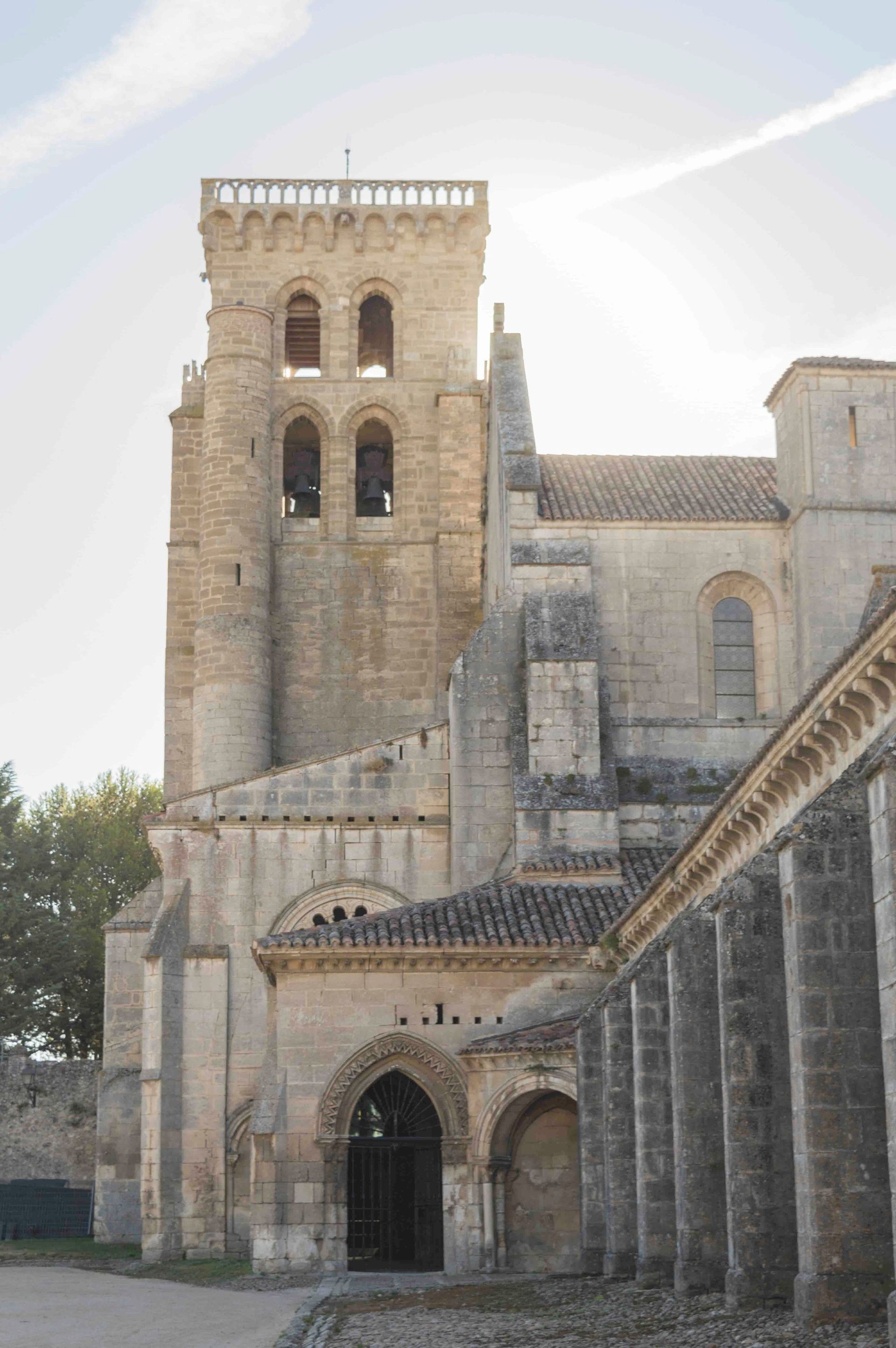 Torre de iglesia de piedra con ventanas arqueadas, campanas y entrada lateral. Luz solar.