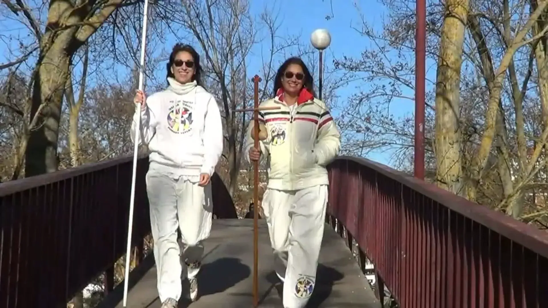 Dos personas caminando sobre un puente, sosteniendo bastones, vistiendo gafas de sol y trajes blancos en un día soleado.