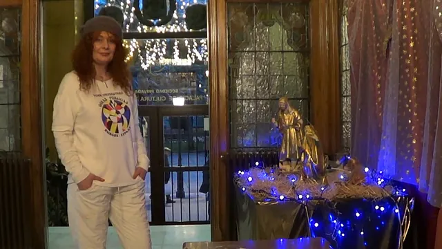 Una mujer vestida de blanco se encuentra cerca de un belén, iluminado con luces azules, en una puerta.