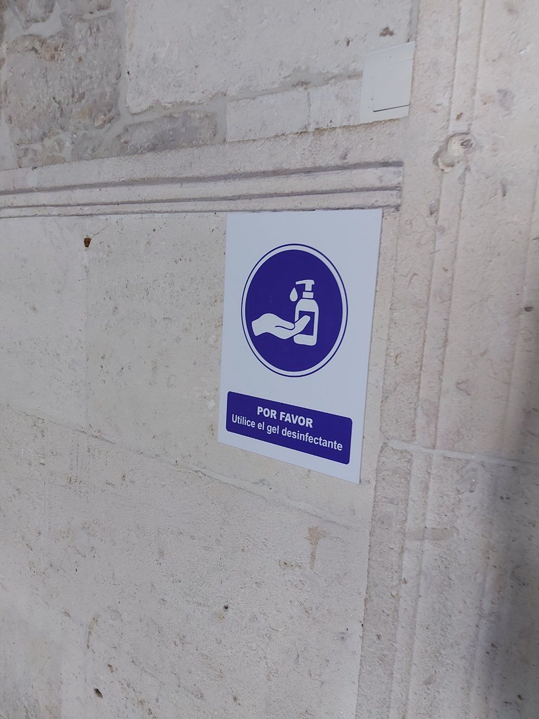 Cartel con un círculo azul que muestra cómo se desinfectan las manos con una botella; sobre una pared de piedra clara.