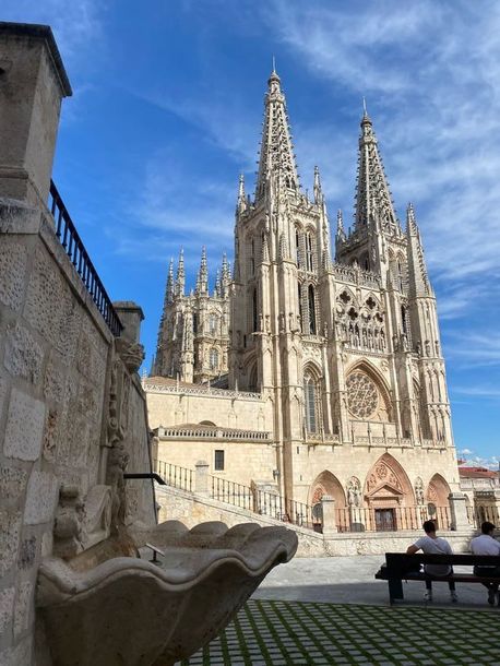 Catedral de Burgos con dos torres bajo un cielo azul; fuente en primer plano, dos personas en un banco.