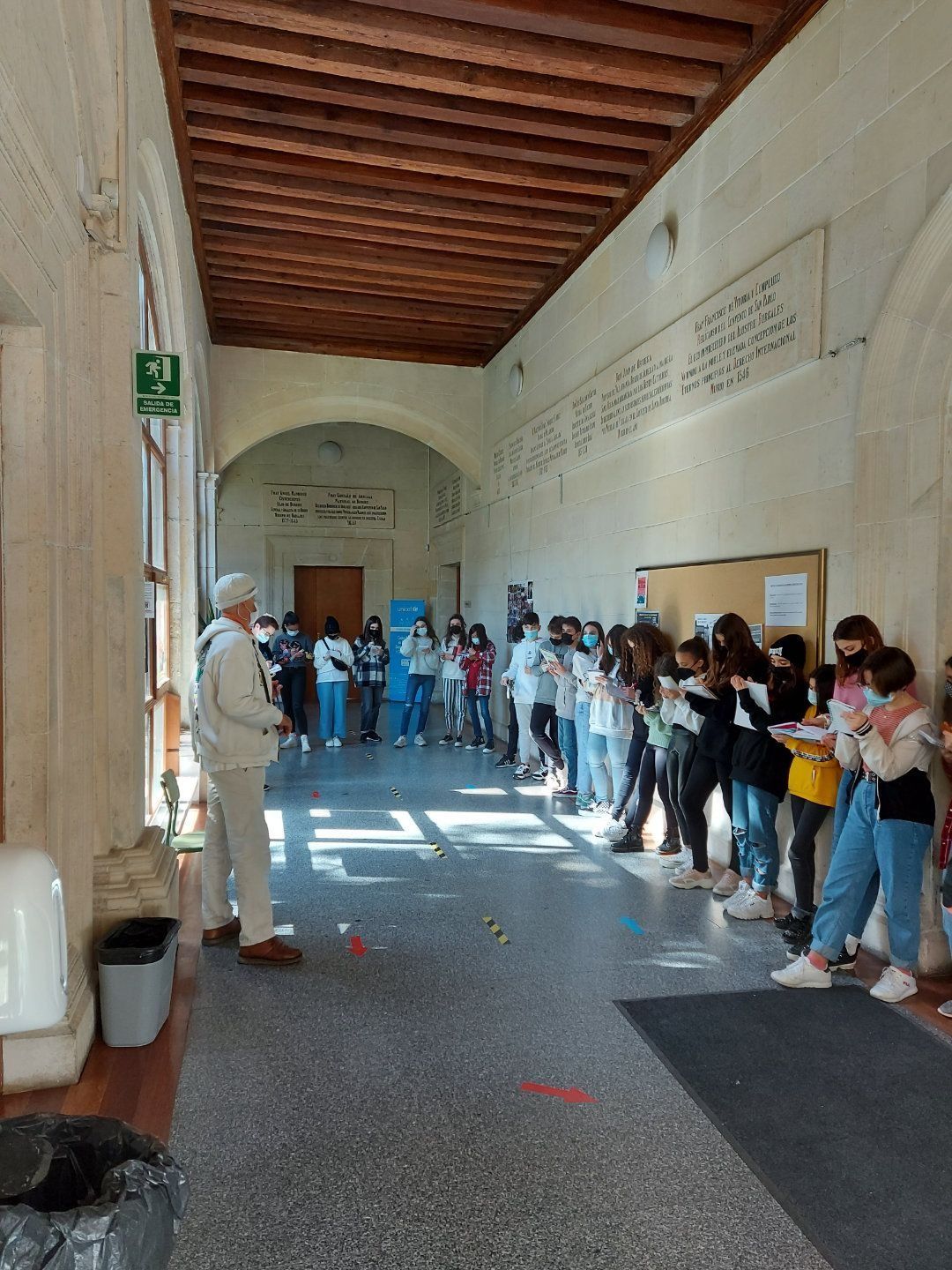 Pasillo de la escuela con estudiantes y profesor, algunos con mascarillas. Los estudiantes hacen fila, algunos con objetos.