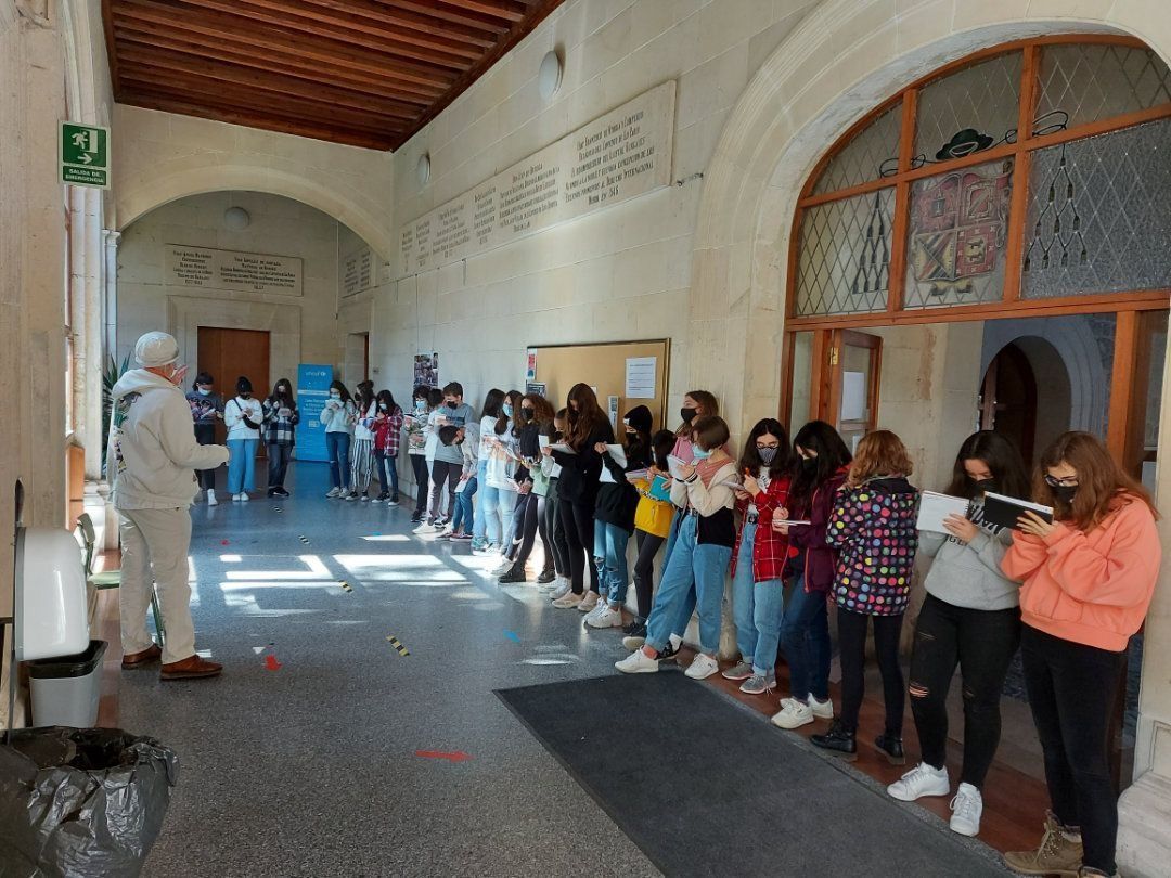 Los estudiantes hacen fila en el pasillo de una escuela, algunos con máscaras, junto a un instructor cerca de una puerta.