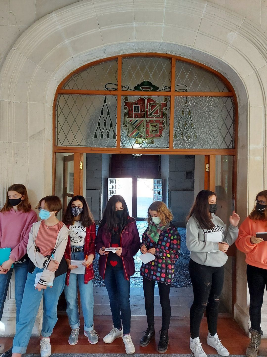 Grupo de chicas enmascaradas frente a una puerta arqueada, sosteniendo papeles. Se ve un escudo de armas sobre la puerta.