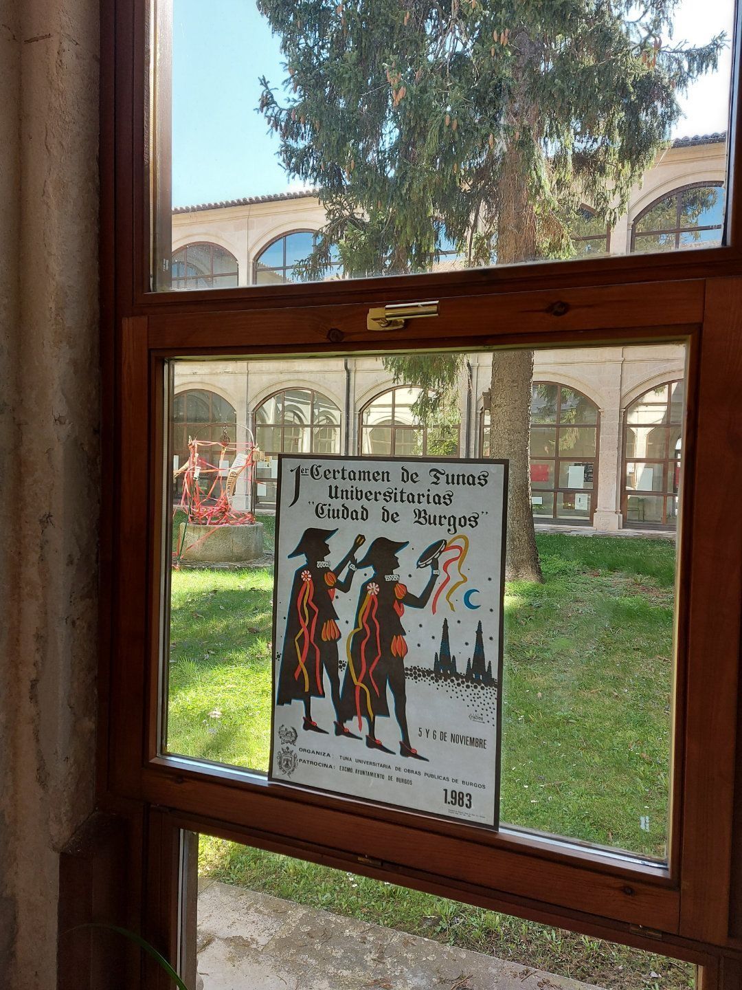 Póster a través de una ventana. Dos siluetas, texto. Edificio al fondo.