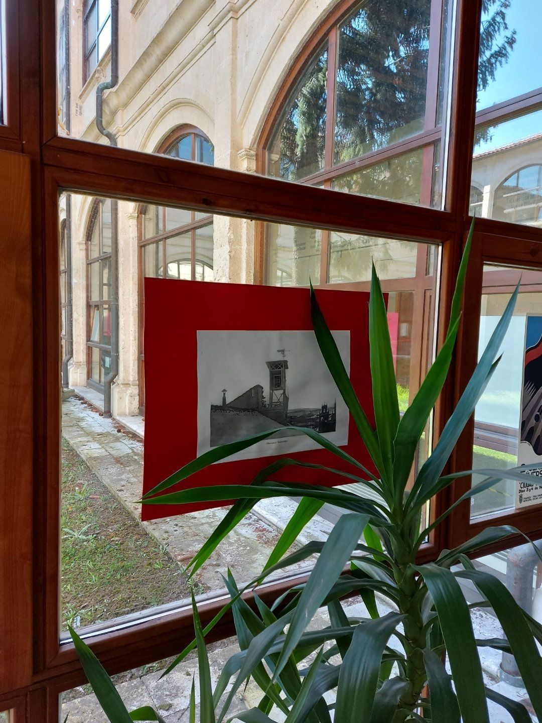 En una ventana con una planta delante se muestra el dibujo de un edificio sobre papel rojo.