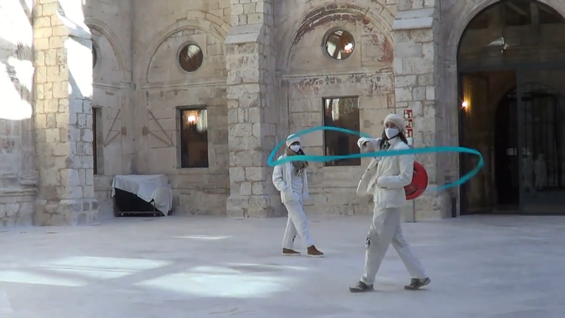 Dos personas vestidas con trajes blancos practican esgrima con una estela de luz azul en un antiguo edificio de piedra.