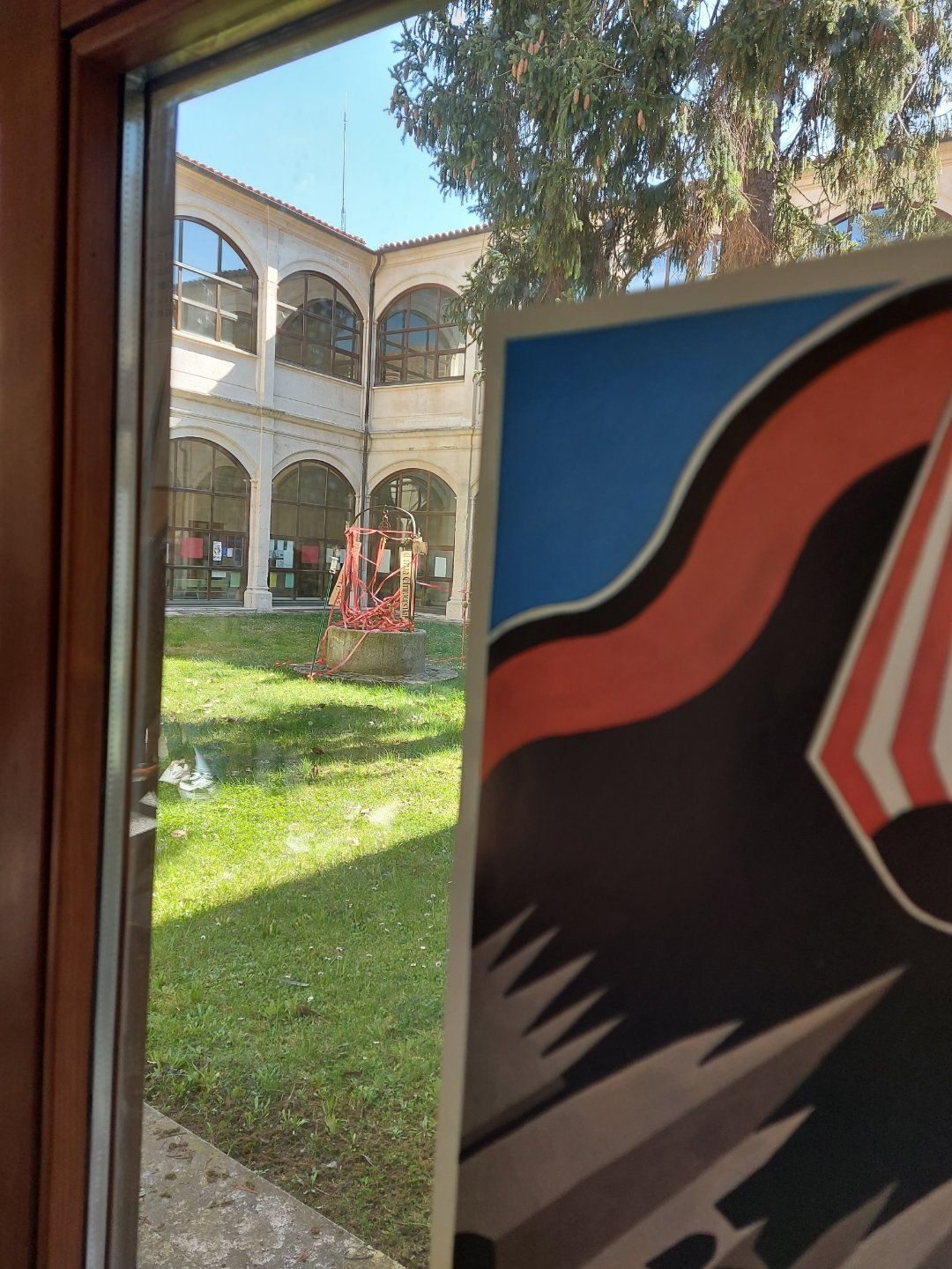 Vista del patio a través de una ventana, parcialmente oculta por un cartel. Césped verde, edificio blanco y cielo azul despejado.