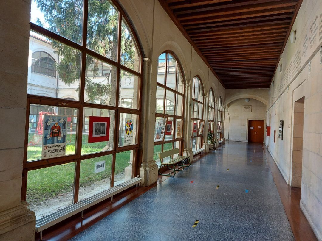 Se ve un largo pasillo con ventanas arqueadas que muestran carteles; un techo de madera, paredes de piedra y una puerta.
