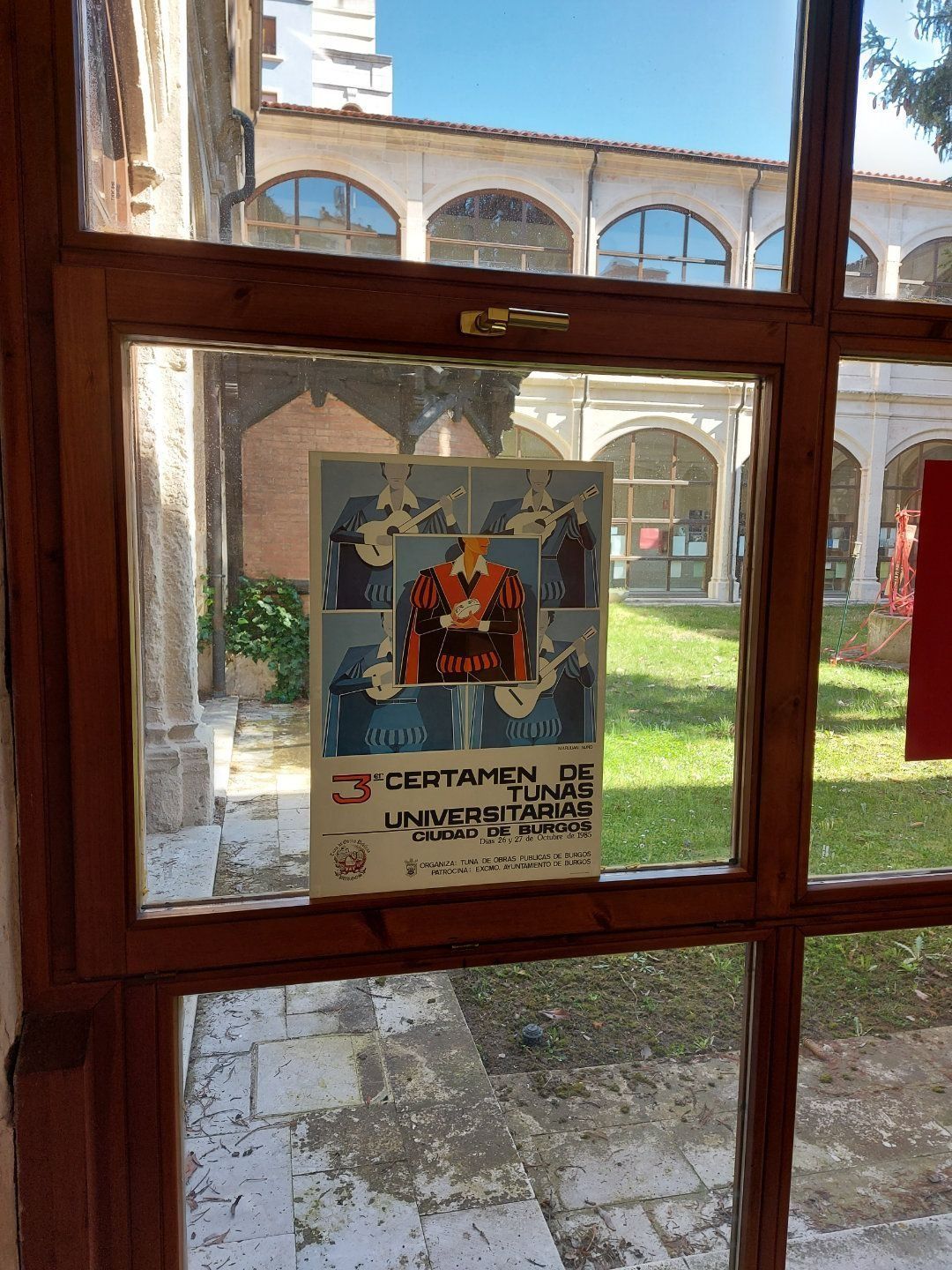 Cartel en un marco de ventana de madera, promocionando un concurso universitario, con un patio de ladrillo visible al fondo.