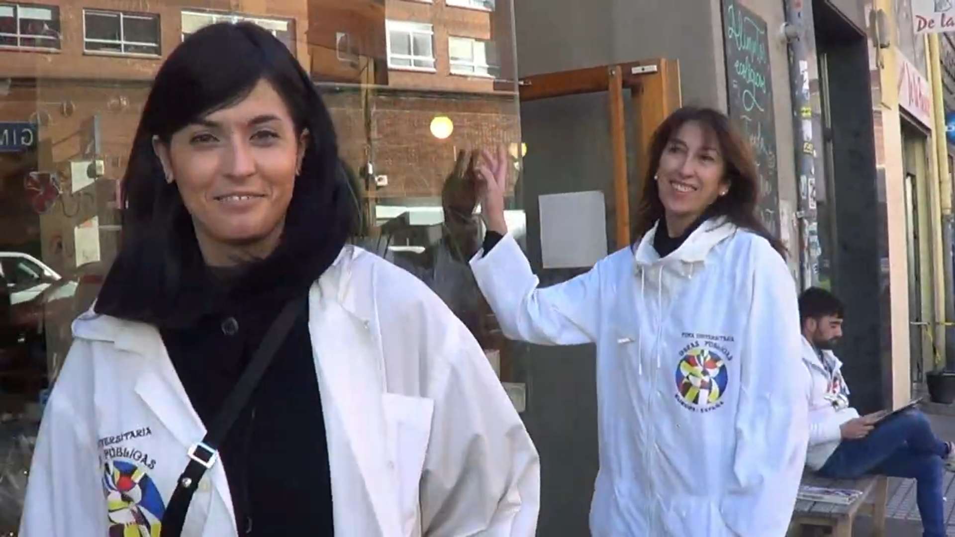 Dos mujeres con batas blancas, sonriendo, frente a una tienda. Un hombre está sentado cerca.