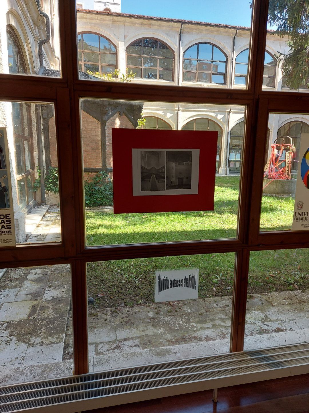 Una fotografía enmarcada por una ventana de madera, que muestra una imagen en blanco y negro sobre un fondo rojo con un césped verde y un edificio al fondo.