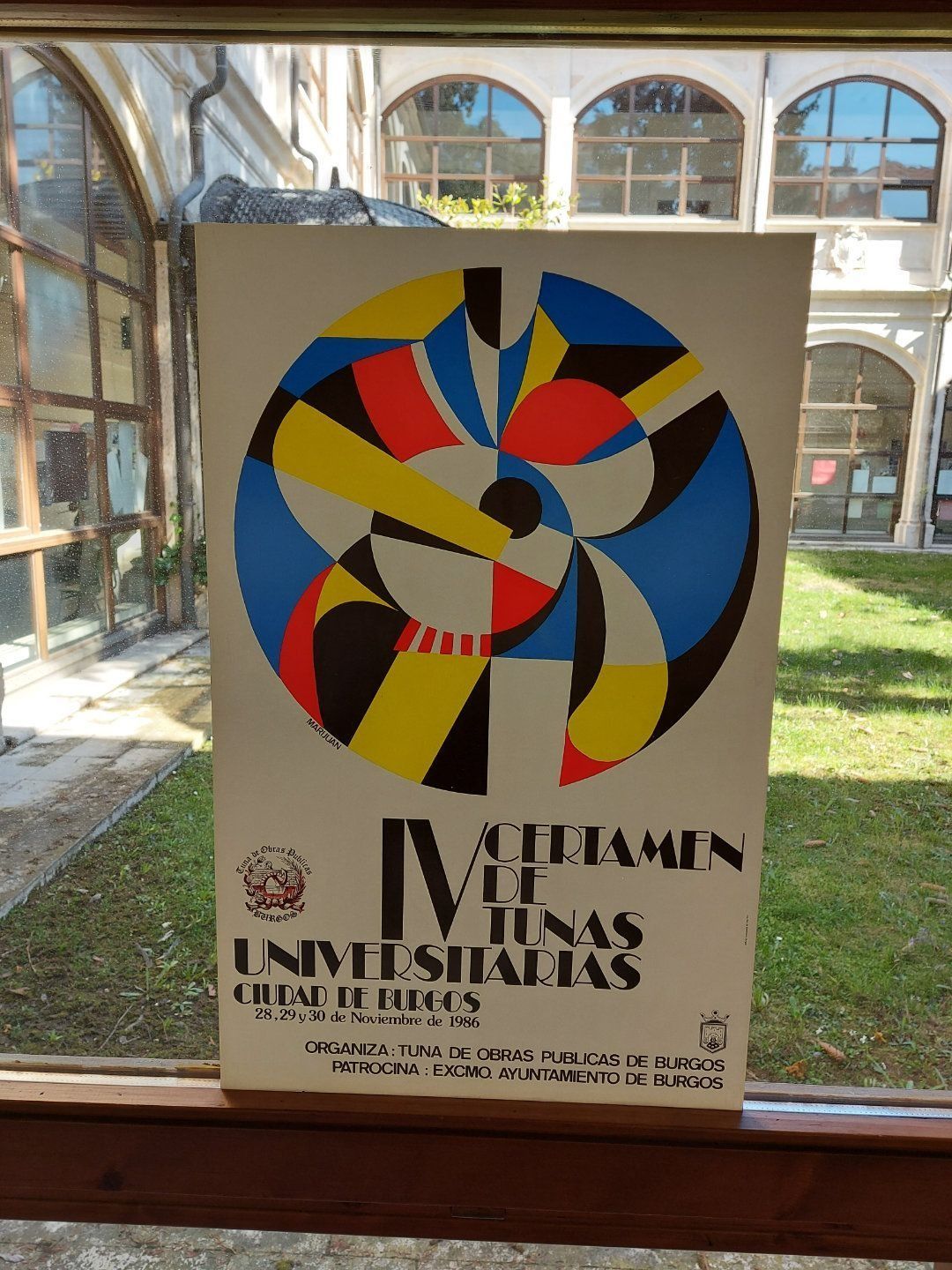 Cartel del IV Certamen de Tunas Universitarias con diseño abstracto colorido, expuesto cerca de una ventana.