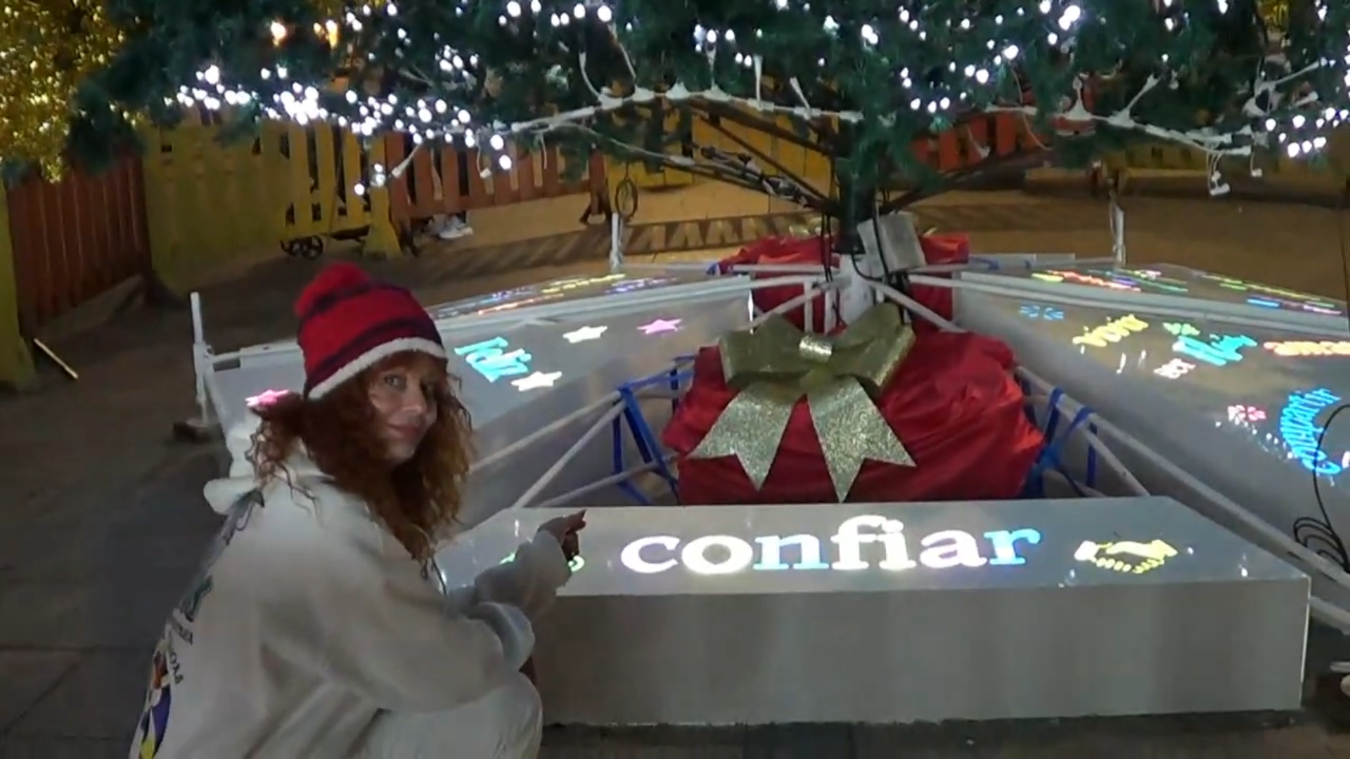 Mujer con gorro de Papá Noel junto a un árbol de Navidad iluminado, con 