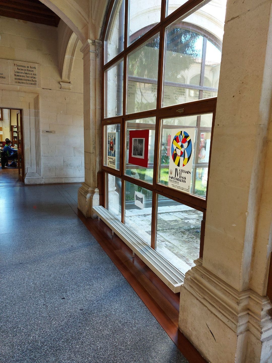Pasillo interior con grandes ventanales que exhiben obras de arte, columnas de piedra y una vista de un jardín al aire libre.