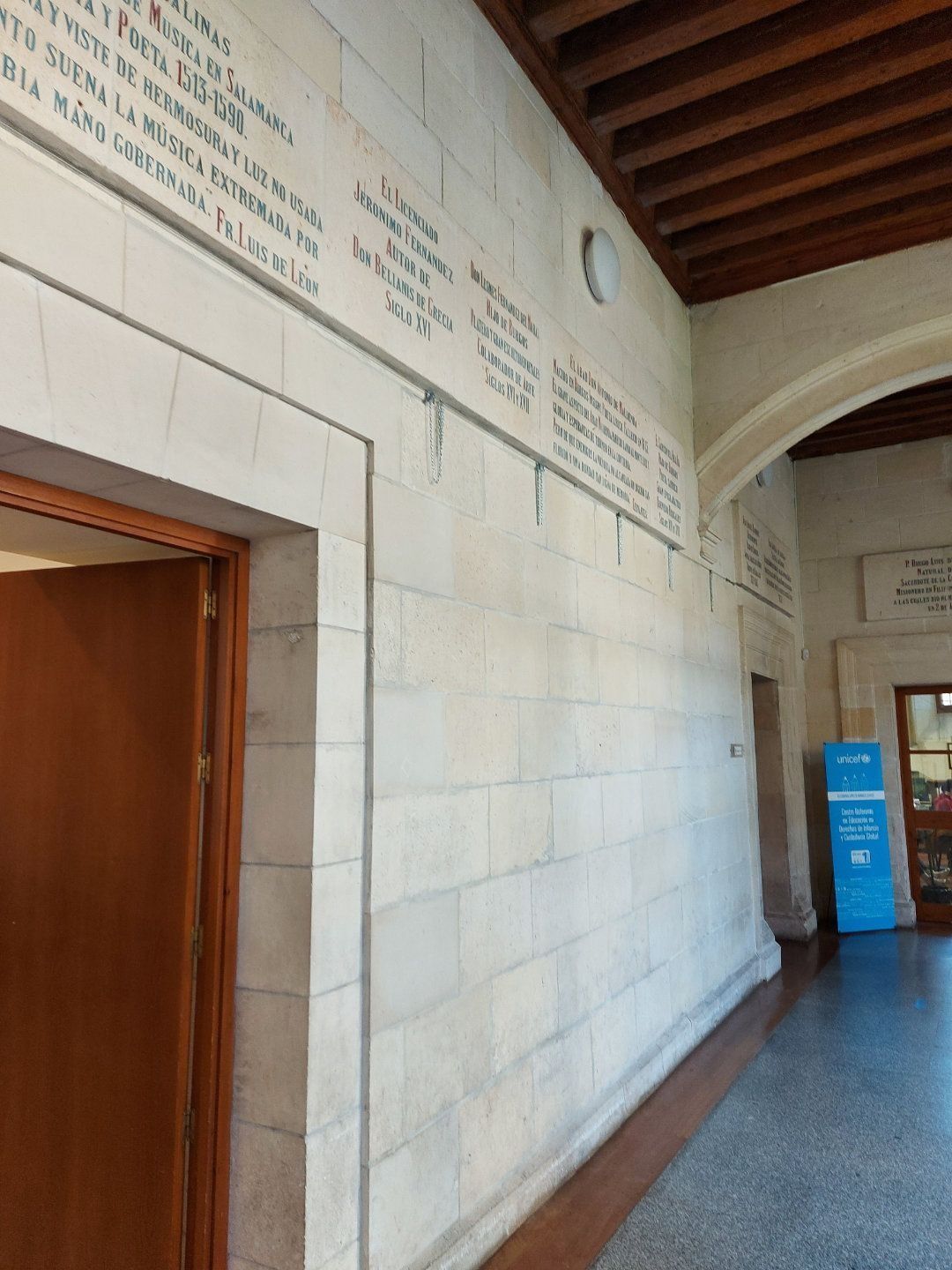 Muro de piedra en un pasillo arqueado, con placas y un marco de puerta de madera a la izquierda.