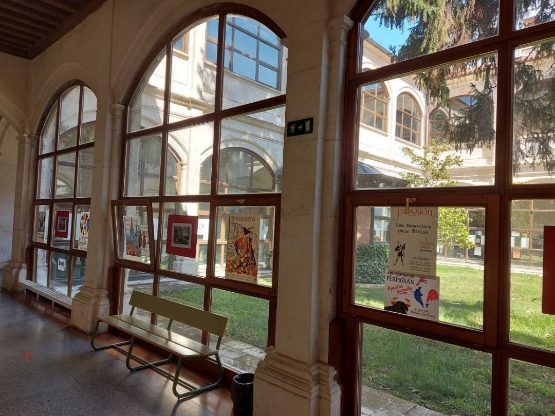 Pasillo con ventanas arqueadas que dan a un patio. Pósteres en la pared, banco a la izquierda. Día soleado.