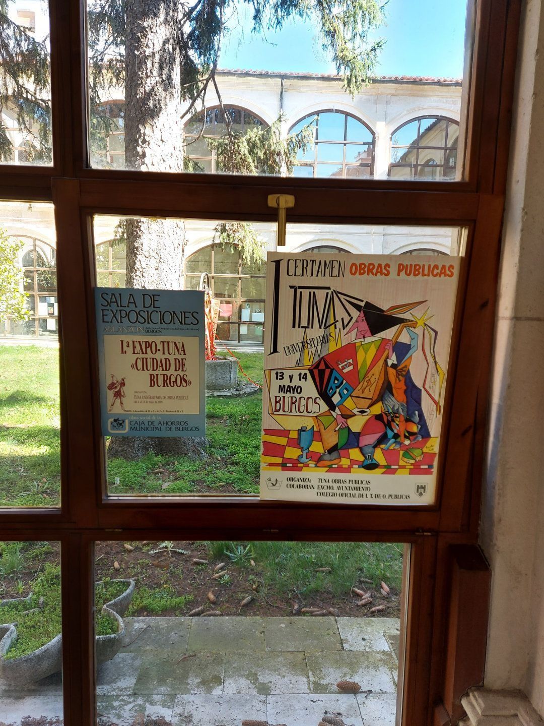 Ventana con dos carteles: uno azul, otro colorido; vista de un patio con árboles.