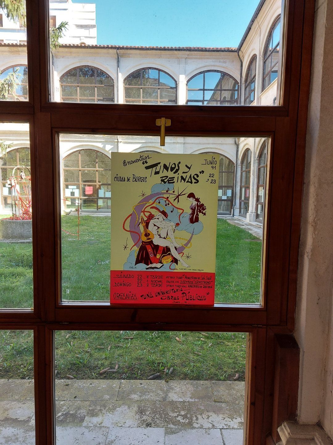 Cartel en ventana: obra de arte colorida de un rostro, texto y un panel inferior rojo; visto a través de una ventana con marco de madera que da a un patio.