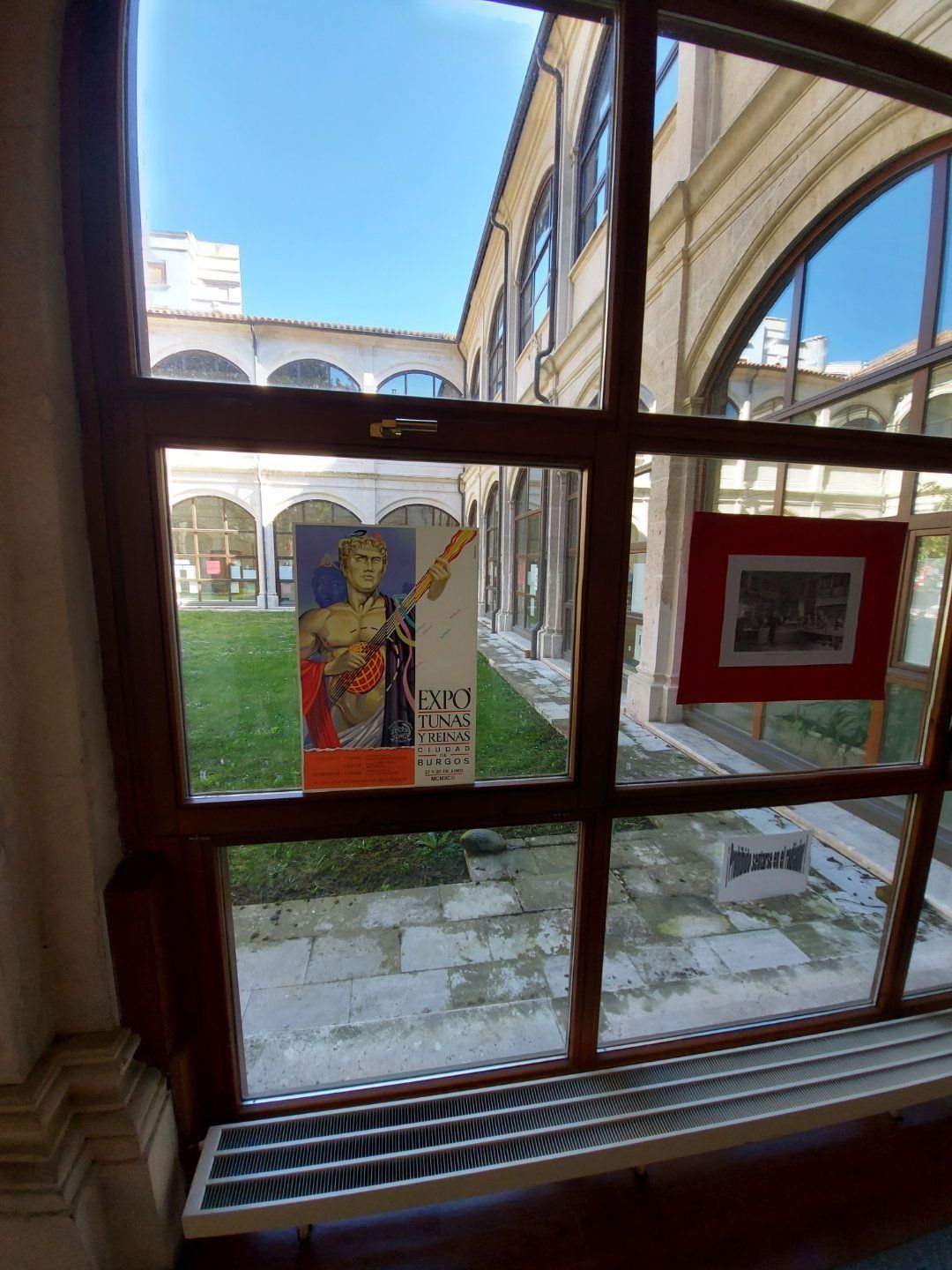 Vista a través de una ventana de un patio interior con carteles y puertas arqueadas en un día soleado.