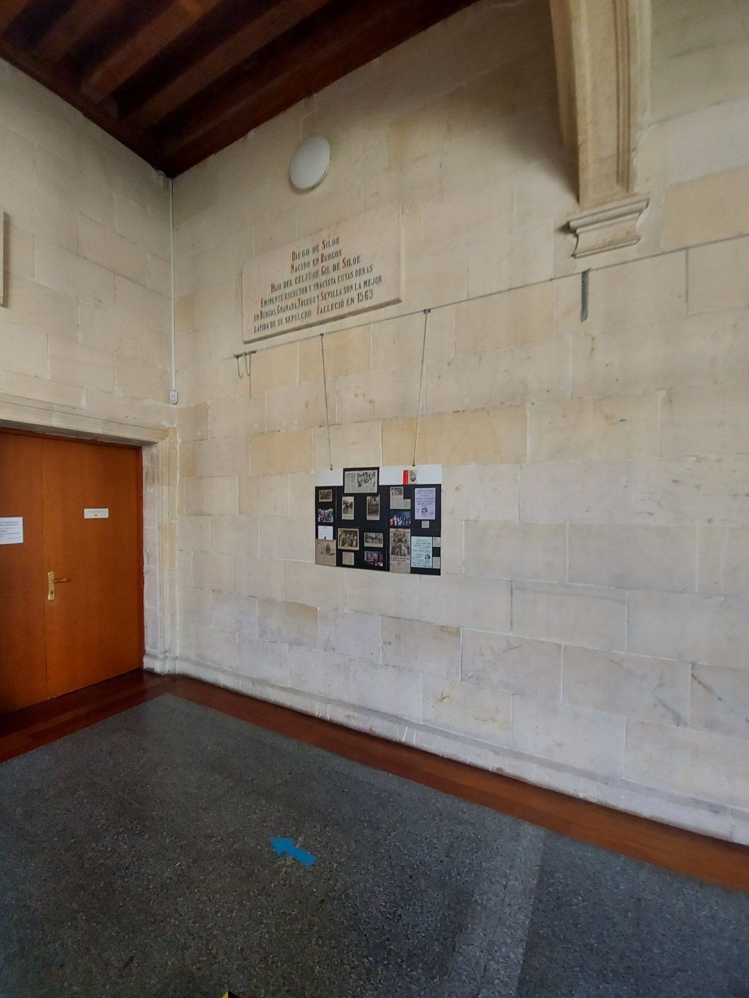 Un muro de piedra dentro de un edificio con una puerta de madera a la izquierda y un tablón de anuncios con papeles en el centro.