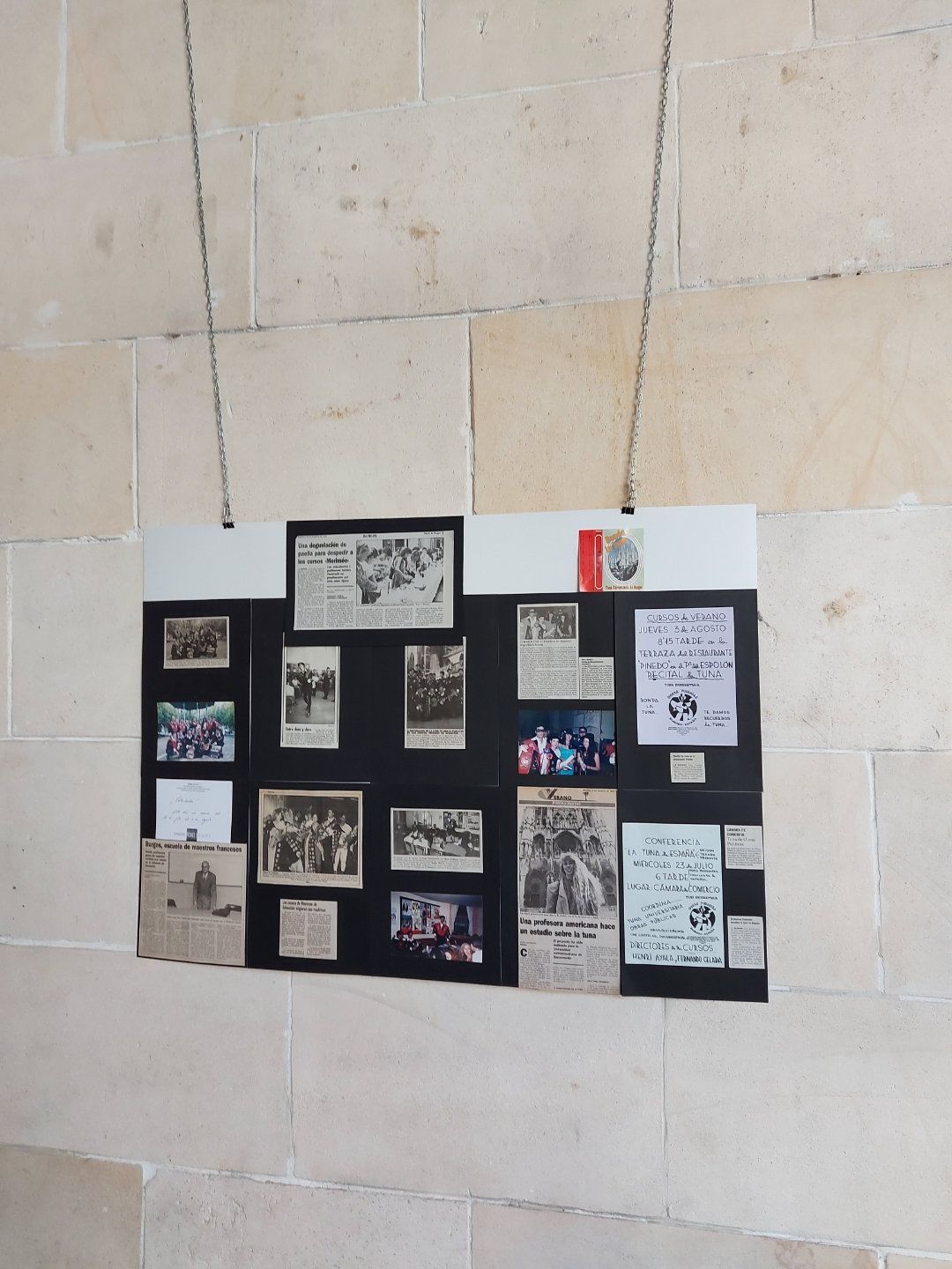 Un tablero negro con fotografías y documentos históricos colgado en una pared de piedra beige claro.