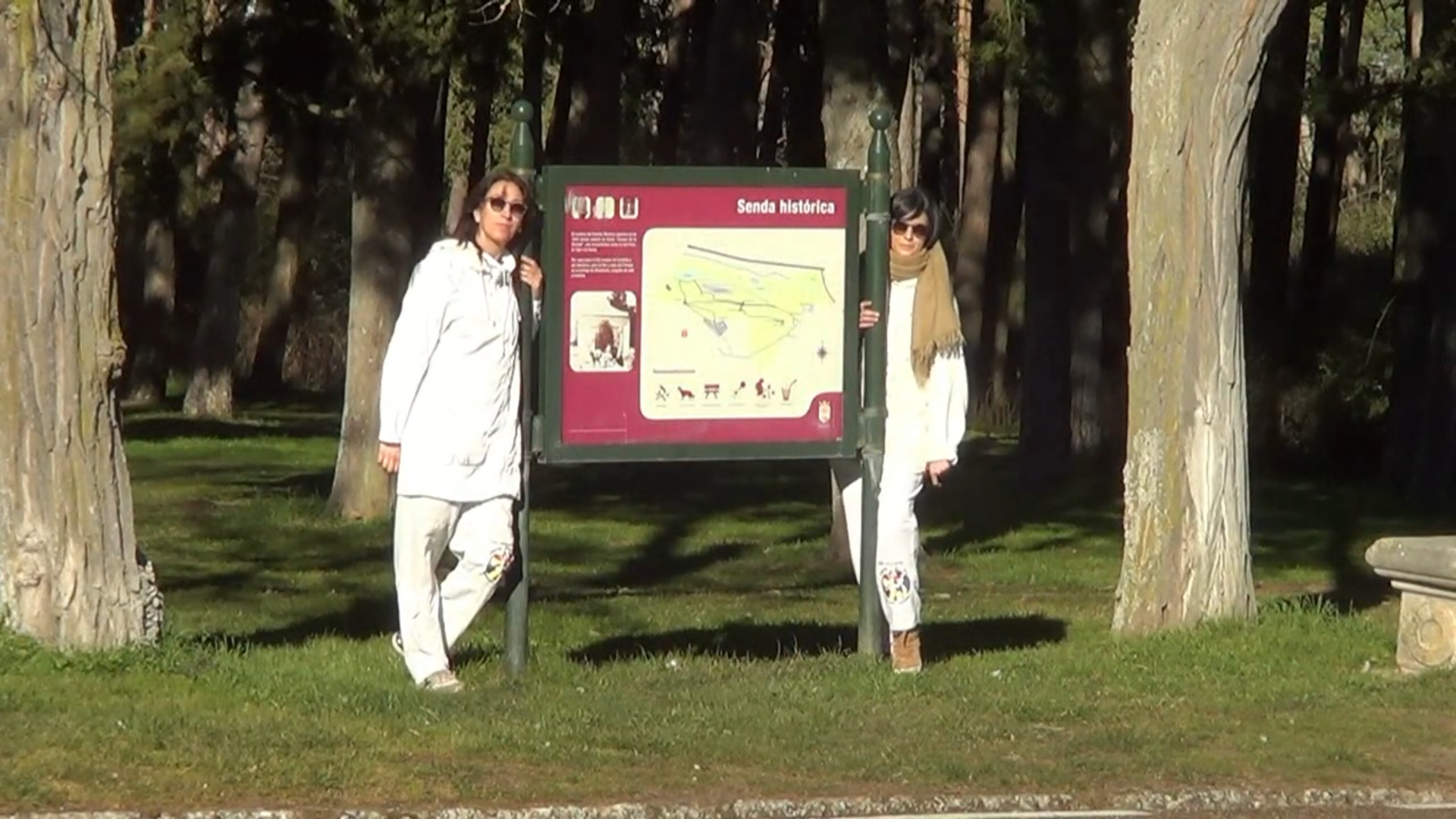 Dos personas vestidas de blanco se encuentran junto a un mapa de un parque en un entorno verde.