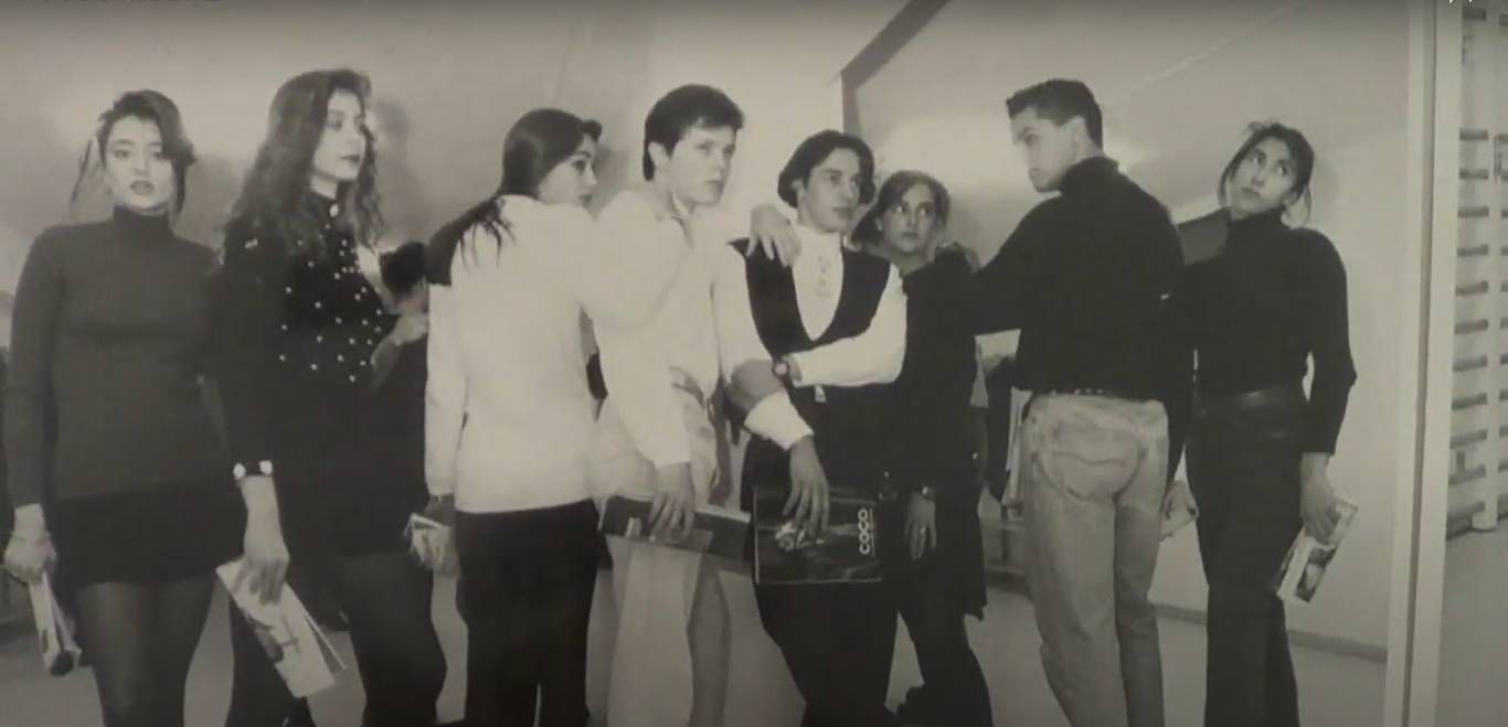 Grupo de jóvenes posando, algunos del brazo, en una habitación. Visten moda de los 90.