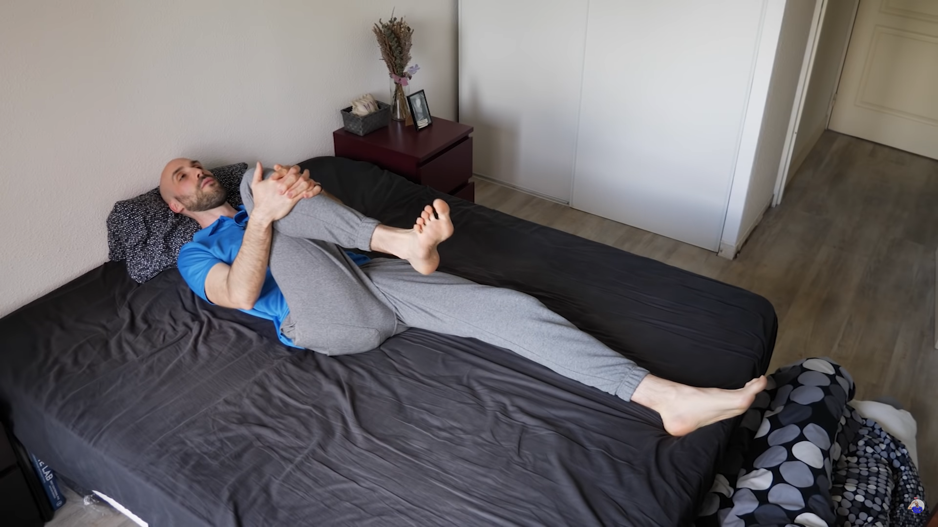 Hombre en la cama, estirando las rodillas hasta el pecho, con pantalones grises y camisa azul. Dormitorio.