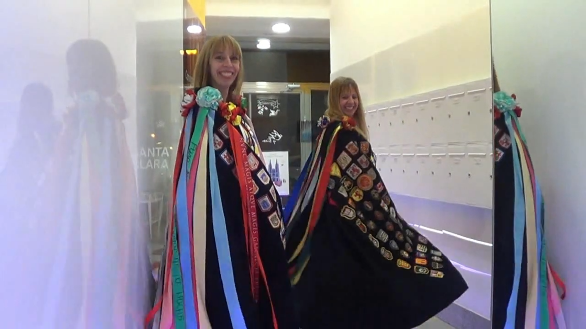 Mujer con capa negra con cintas y parches de colores, sonriendo cerca de un espejo en un pasillo.